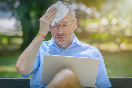 额头出汗人在公园里使用笔记本电脑和用纸巾擦拭额头时会中暑.照片