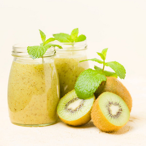 在黄色柔和的背景上用新鲜的绿色薄荷叶和原始成熟的切果装饰的猕猴桃奶昔透明的混合维生素鸡尾酒罐, 用于健康食品概念