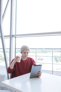 一个微笑的年轻男子的肖像, 穿着时髦的连衣裙, 坐在咖啡馆里, 手里拿着一杯咖啡和一块药片。学生使用互联网在一个光咖啡馆的平板电