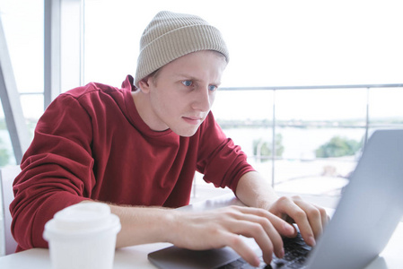 严肃的年轻人穿着休闲服, 坐在桌边, 在笔记本电脑键盘上打字。年轻的自由职业者在一个小咖啡馆的笔记本电脑工作。博主在电脑上写文章