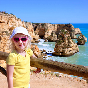 一位游客, 戴着帽子和太阳镜的小女孩, 欣赏葡萄牙南部的 Marinha 海滩上美丽的悬崖上的美景。家庭徒步旅行在拉各斯最壮观的