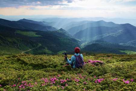 在杜鹃花的草坪上, 旅游女孩坐在后袋里。景观与伟大的美丽的山在太阳光线。生态旅游。美丽的夏日风光