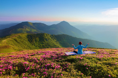 多彩的 karemat。在莲花姿势的瑜伽女孩。草地上有杜鹃花花。高山。神奇的森林。冥想。放松。夏天的风景。温暖的早晨太阳光线