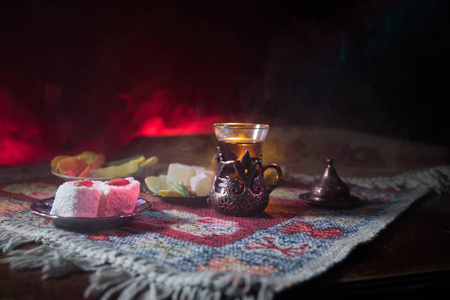 阿拉伯茶在玻璃与东方小吃在地毯上的黑暗背景与灯光和烟雾。东方茶的概念。空的空间。选择性聚焦