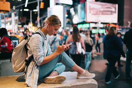 时髦女孩坐在时代广场与智能手机发送照片从地标在晚上, 女性旅行者在社交网络上通过手机和互联网连接的旅游观光在新的你