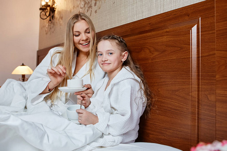 微笑的母亲和孩子在床上吃早饭, 在舒适的旅馆房间里。家庭爱关怀的概念