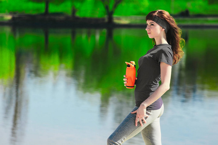 在运动服的女孩拿着一瓶水，视线和微笑，站在沙滩上后，锻炼