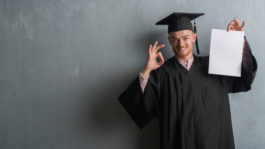 灰色垃圾墙上的年轻红头发的人穿着研究生制服持有学位做 ok 标志与手指, 优秀的标志