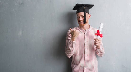 灰色垃圾墙上的年轻红头发的男子戴着研究生帽持有学位指向和显示与拇指到一边, 快乐的脸微笑着