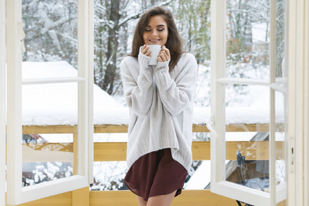 快乐的女人与一杯热饮料站在阳台上的雪冬日