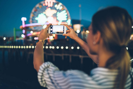 后视图的妇女访问旅游剧院在晚上拍照摩天轮的智能手机, 时髦的女孩使用蜂窝相机, 以制作圣莫尼卡码头景点的形象在临近