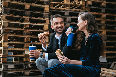 微笑情侣在豪华服装与热狗和苏打饮料坐在街上的木托盘侧面视图