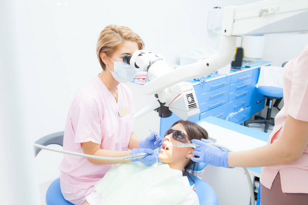年轻的女牙医治疗根管显微镜在牙科诊所。年轻女病人躺在牙医的椅子上，张开嘴。戴着口罩和手套的牙医