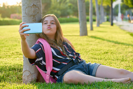金发的学生女孩与智能手机在公园回学校坐在草地上