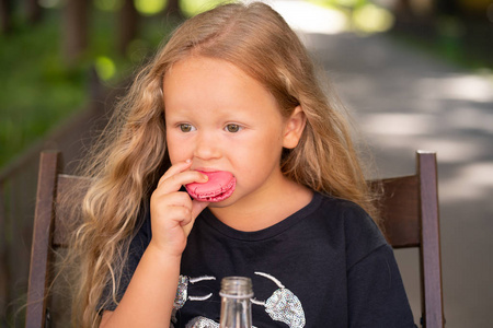 可爱的高加索小女孩坐在木椅上, 吃着甜甜的粉红色玛卡龙独自一人户外