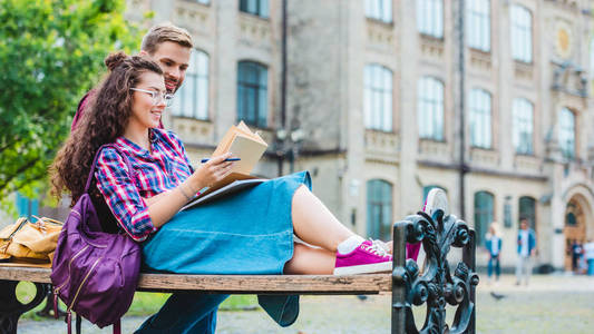 微笑的年轻妇女与书和笔记本依靠男朋友在公园的木凳上