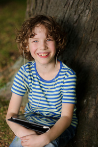 开朗的男孩坐在一棵树下与平板电脑在腿上