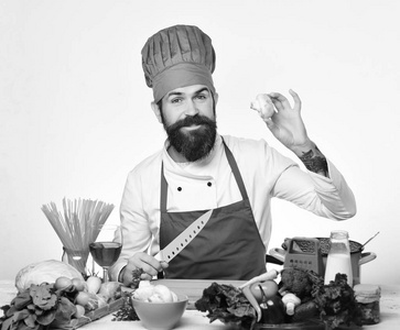 有胡子的男人在白色的背景下拿着蘑菇和刀子。厨师与微笑在勃艮第制服坐厨房桌
