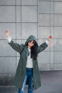 好漂亮的年轻时髦女人摆在太阳镜户外时尚特写肖像。布鲁内特快乐的女孩，穿着绿色雨衣和粉红色运动鞋走城里的街上