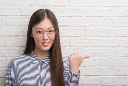 年轻的中国企业妇女在砖墙指向和显示与拇指到边与愉快的面孔微笑