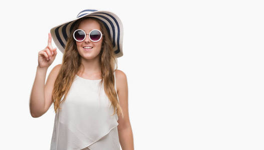戴着太阳镜和夏日帽子的年轻金发女郎惊讶于一个想法或问题, 手指与快乐的脸, 第一