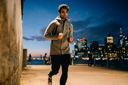 积极的男性慢跑者身着灰色运动服运行在城市设置和听音乐播放器在夜间