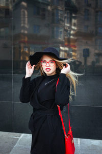 美丽的年轻女子在黑色大衣和帽子，穿着红色眼镜和一个红色的书包，走在这个城市。反映在窗口中城市