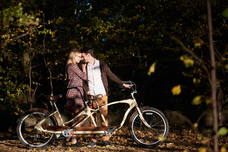 年轻幸福浪漫的情侣, 胡子男子和迷人的时尚女子紧密结合在一起在秋季公园或森林模糊的黑暗树木茂密的树叶背景上的双联自行车户外