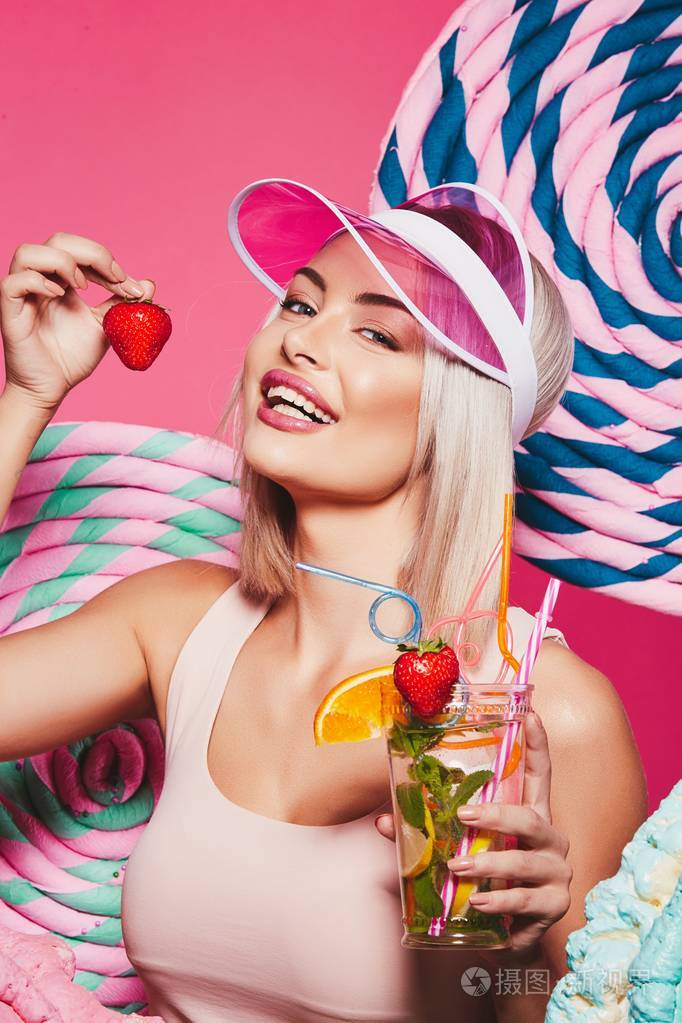 金色的头发穿着顶部和粉红色的帽子站在粉红色背景举行柠檬水和吃草莓