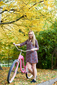 苗条的快乐金发美女在眼镜和短礼服与粉红色夫人自行车站在高大的树上, 美丽的绿色和金色的秋天叶子阳光明媚的背景