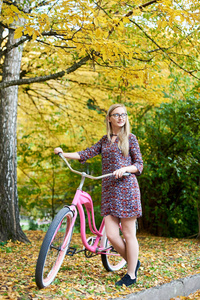 微笑的金发女郎时尚迷人的眼镜和短礼服与粉红色的女士自行车独自站在高大的树下, 享受美丽的绿色和金色的秋天树叶在晚上