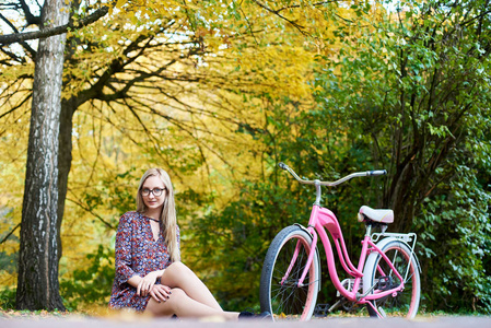 金发女郎在眼镜和时髦的短礼服坐在地面上粉红色夫人自行车下高大的树与金色的叶子在美丽的五颜六色的秋天树叶阳光明媚的背景