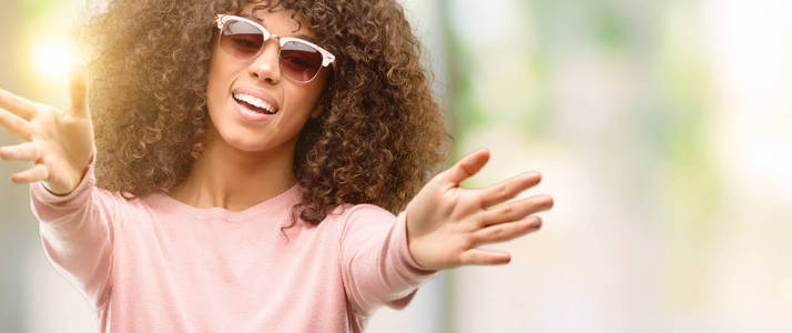 身穿粉红色太阳镜的非洲裔美国妇女看着相机张开双臂微笑着拥抱。快乐的表达拥抱幸福