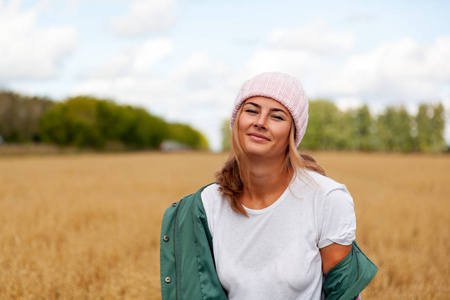 年轻的黑发妇女在一个针织粉红色的帽子, 从自然的毛皮微笑, 并提出积极的秋季一天, 对背景的黄金场和森林。村庄 Livestyl