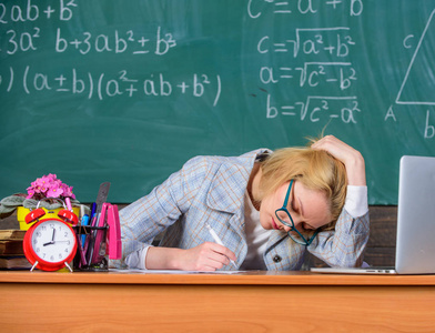 女教师坐桌教室黑板背景。还在工作工作远远超过实际的学校一天。老师疲倦的面孔在课以后继续工作。教师忙于文书工作和研究