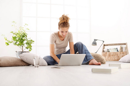 年轻女子与电脑, 智能手机和书籍, 坐在地板上的客厅在白色的宽窗口背景