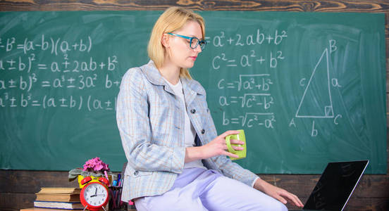 休息一下。未来教师必须考虑的工作条件。女镇定老师拿着杯子喝坐桌教室黑板背景。教师工作条件