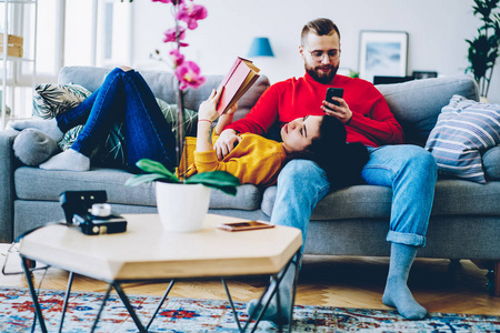 集中的人发送短信在智能手机上使用家庭 wifi 在闲暇的时候, 他的女朋友在沙发上阅读有趣的书, 情侣在舒适的公寓内饰一起休息