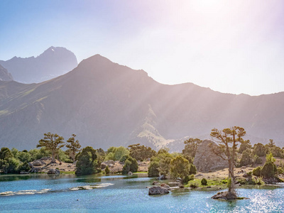 山湖与蓝色水和一棵树在阳光下在岩石山背景。文芳山脉, 塔吉克斯坦, 中亚