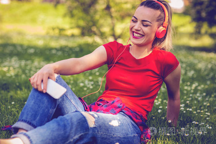 美丽的快乐女孩与耳机和智能手机听音乐在草地上公园