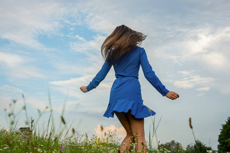 美丽的微笑的年轻女子是跳舞和旋风在一个蓝色的礼服在草地上的花朵背景在夏季