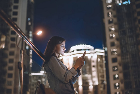 有吸引力的妇女的侧面视图使用智能手机在街道与夜城市灯背景