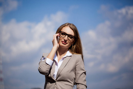 美丽的女企业家站在天空背景，谈电话的肖像。女人穿着西装和眼镜