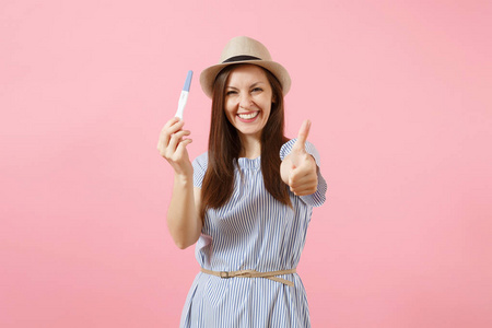 兴奋的快乐女人穿着蓝色的连衣裙, 帽子握在手里, 看着怀孕测试被隔离在粉红色的背景上。医疗保健妇科孕产妇生育的概念。复制空间