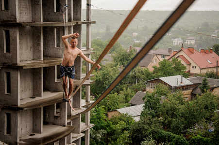 年轻无畏的人走在slackline和举起手对空的建筑和树在雨天