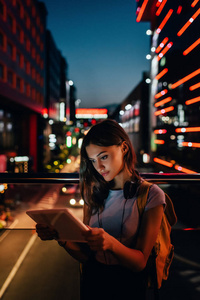 带耳机的年轻妇女的肖像在街道上用平板电脑与夜间城市灯光背景