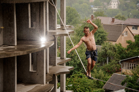 积极无畏的人走在一个slackline和举起手对空建筑和树木在雨天