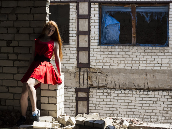 红头发薄的苗条的女孩穿着红色背景被毁的建筑物墙上的雀斑