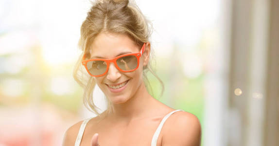 年轻漂亮的女人戴着红色的太阳镜微笑着广泛显示大拇指向上的手势相机, 表达的喜欢和批准