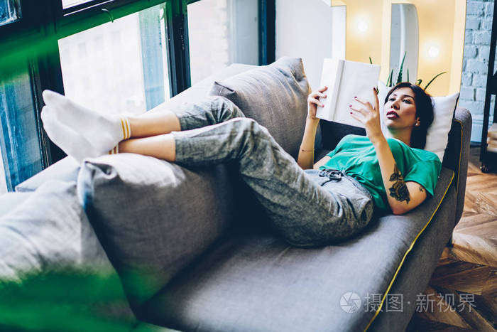 聪明的时髦女孩躺在舒适的沙发上, 阅读感兴趣的书花娱乐时间在家里最